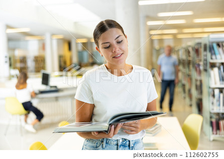 Carefree young lady reading the magazine and smiling while standing in library Carefree young lady reading the magazine and smiling while standing in library 112602755