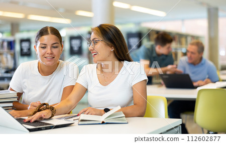 Two young women laughing and speaking loudly while using laptop in the library 112602877