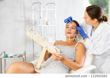 Adult female cosmetologist examining face of female patient 112602882