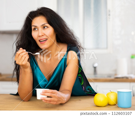 Woman standing at kitchen table and eating yoghurt Woman standing at kitchen table and eating yoghurt 112603107
