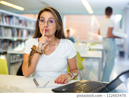 Serious young Latin lady bringing the index finger to lips and looking at the camera working on computer in the library Serious young Latin lady bringing the index finger to lips and looking at the camera working on computer in the library 112603470