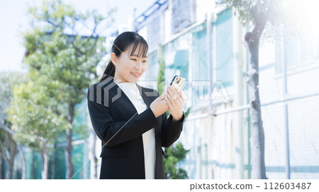 一位身穿西裝的年輕日本（亞洲）女性在新鮮的綠色植物中看著她的智慧型手機。 112603487