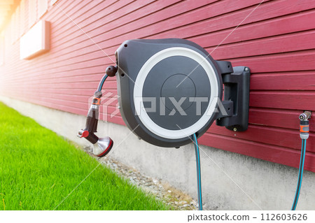 Wall Mounted Retractable Hose Reel. Modern garden lawn watering tip hose with sprinkler hanged at home wall. Lawn and plant tree smart irrigation water system equipment tool 112603626