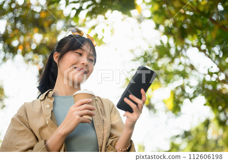 Satisfied Asian woman drinking coffee from paper cup using mobile phone in the park 112606198