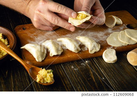 The chef is preparing pelmeni on the kitchen table with dough and potatoes. Concept of preparing a delicious lunch with dumplings The chef is preparing pelmeni on the kitchen table with dough and potatoes. Concept of preparing a delicious lunch with dumplings 112609068