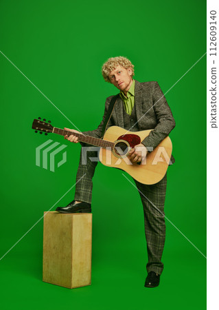 Curly young man in a suit playing guitar against green studio background. Poster for live music event Curly young man in a suit playing guitar against green studio background. Poster for live music event 112609140