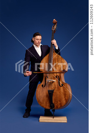 Stylish man in suit paying double bass, making classical music live performance against dark blue background 112609143