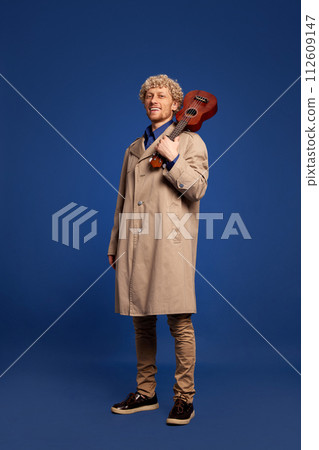 Smiling man in stylish trench coat standing with ukulele against dark blue background. Creative solo performance Smiling man in stylish trench coat standing with ukulele against dark blue background. Creative solo performance 112609147