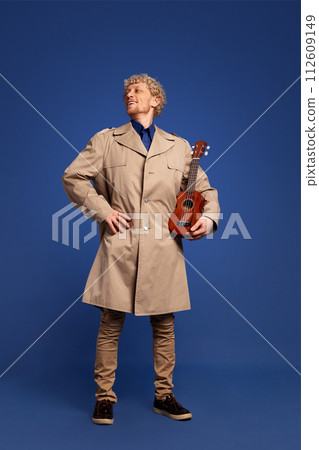 Smiling man in stylish trench coat standing with ukulele against dark blue background. Creative solo performance Smiling man in stylish trench coat standing with ukulele against dark blue background. Creative solo performance 112609149