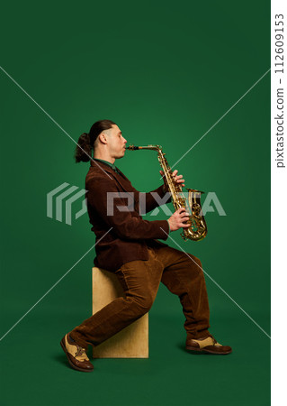 Talented and artistic young man, musician in costume sitting and playing saxophone against dark green background. Live concert 112609153