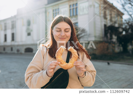 Attractive young female 30s tourist with backpack is holding bagel, traditional polish snack in Krakow. Traveling Europe in spring. Selective focus. High quality photo Attractive young female 30s tourist with backpack is holding bagel, traditional polish snack in Krakow. Traveling Europe in spring. Selective focus. High quality photo 112609742