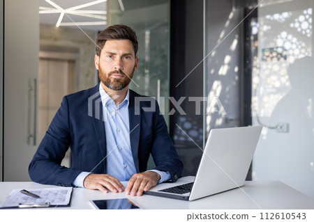 Portrait of a successful and confident young man lawyer in a business suit sitting at an office desk, working with a laptop and documents, looking seriously at the camera. Portrait of a successful and confident young man lawyer in a business suit sitting at an office desk, working with a laptop and documents, looking seriously at the camera. 112610543