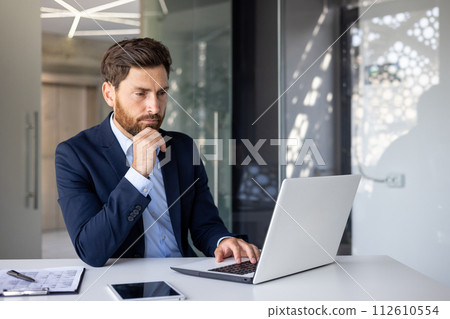 A serious and thoughtful young man businessman is sitting in the office at the desk and is concentrating on his laptop. 112610554