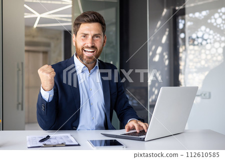 Portrait of a happy young businessman showing a hand gesture of success and victory to the camera. He sits in the office at the table and works on a laptop and with a project. 112610585