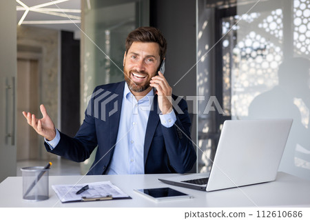 Portrait of a successful and smiling young man businessman, salesman, broker emotionally talking on the phone with clients, sitting at the desk in the office and looking at the camera. 112610686