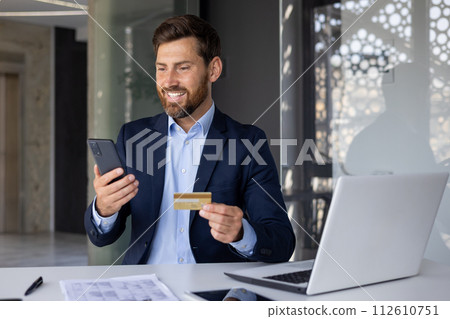 Successful financial transaction and growth. A smiling and satisfied young businessman is sitting at the desk in the office, holding a bank credit card and looking at the phone screen. 112610751
