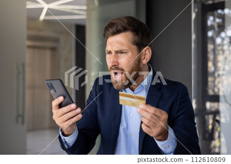 Close-up photo of angry young businessman in suit standing in office, holding golden credit card in hand and looking disappointedly at mobile phone screen. 112610809