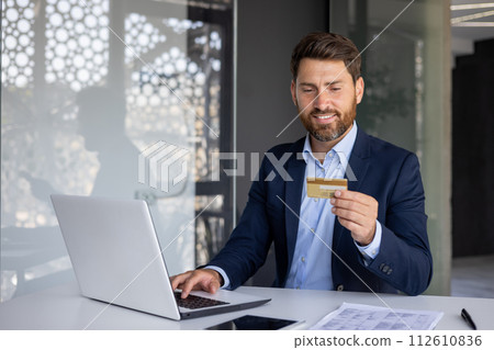 A smiling young man works in the office at a laptop, types on a keyboard and uses a credit card in his hands, conducts online transactions, checks an account. 112610836