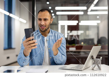 A young African American man works in the office sitting worried at the desk and looking at the phone screen in frustration and shock. A young African American man works in the office sitting worried at the desk and looking at the phone screen in frustration and shock. 112610922