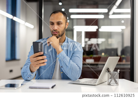 Serious young hispanic man sitting in the office at the desk and looking thoughtfully at the phone screen with his hand on his chin. Serious young hispanic man sitting in the office at the desk and looking thoughtfully at the phone screen with his hand on his chin. 112610925