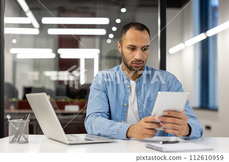 A serious Latin American man in a blue shirt works in a modern office, sits at a desk with a laptop and concentrates on using a tablet in his hands. 112610959