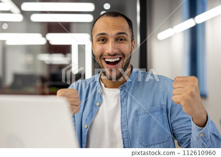 Close-up portrait of a happy young hispanic man sitting in a modern office, in front of a laptop and looking at the camera with a victory gesture. 112610960