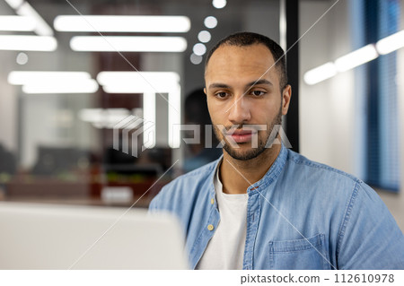 Close-up photo of a young hispanic man in a blue denim shirt, sitting at a desk in an office and looking seriously at a laptop screen. Close-up photo of a young hispanic man in a blue denim shirt, sitting at a desk in an office and looking seriously at a laptop screen. 112610978