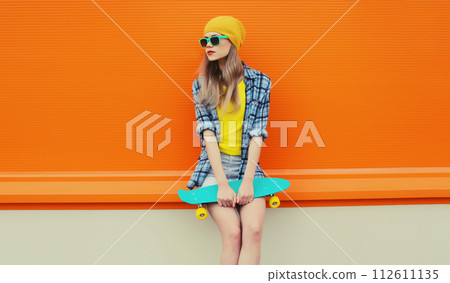 Summer portrait of stylish young woman with skateboard in colorful clothes on orange background Summer portrait of stylish young woman with skateboard in colorful clothes on orange background 112611135