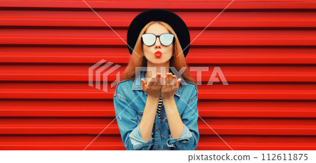 Stylish young woman model blowing kiss in jean jacket, black round hat on red background Stylish young woman model blowing kiss in jean jacket, black round hat on red background 112611875