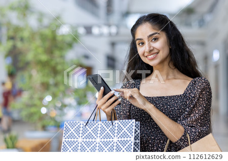Close-up portrait of a young Indian girl standing in a supermarket with shopping bags, holding a mobile phone, smiling and looking at the camera. Close-up portrait of a young Indian girl standing in a supermarket with shopping bags, holding a mobile phone, smiling and looking at the camera. 112612629