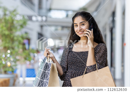 Indian young smiling woman standing in a shopping mall, holding paper shopping bags, talking on the phone and looking at the camera. 112612630