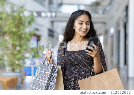 A happy young Indian woman is shopping in a shopping center, standing inside, holding paper bags with goods and using a mobile phone. 112612632