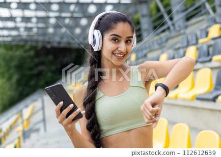 Close-up portrait of a young Indian woman in headphones doing sports, standing in a stadium, holding a phone in her hand and using a smart phone tracker on her wrist, smiling at the camera. 112612639
