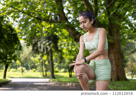 Indian young girl standing in a park in a tracksuit and holding her hands on her knee, feeling a lot of pain, sprained her muscles after running and doing sports. 112612658