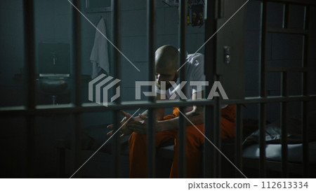 Criminal in orange uniform sits on bed in correctional facility Criminal in orange uniform sits on bed in correctional facility 112613334