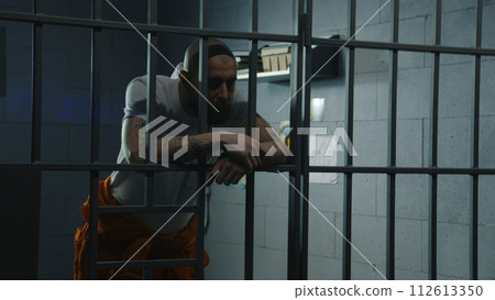 Inmate stands in prison cell leaning and holding hands on metal bars Inmate stands in prison cell leaning and holding hands on metal bars 112613350