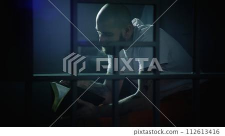 Male prisoner sits on bed in prison cell, reads Bible 112613416