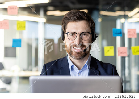 Professional male with beard and glasses wearing a headset and a suit, working on a laptop in a modern office environment. Professional male with beard and glasses wearing a headset and a suit, working on a laptop in a modern office environment. 112613625