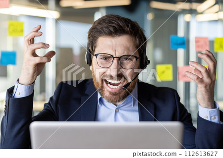 An overwhelmed businessman in a headset shows frustration while working at his desk filled with sticky notes in a modern office. 112613627