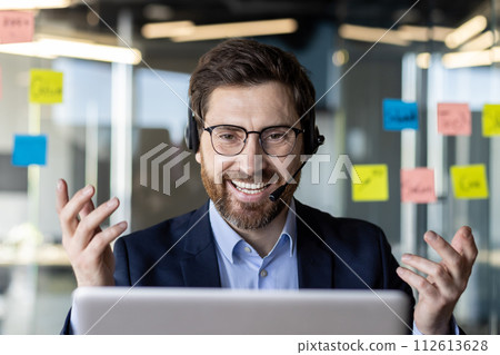 A happy, bearded businessman in a blue suit using a laptop. He wears a headset, gesturing during a video call in a modern office with sticky notes on the glass. 112613628