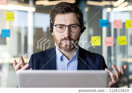 Focused male professional using a headset during a video conference, with sticky notes on the glass behind him in a modern office setting. Focused male professional using a headset during a video conference, with sticky notes on the glass behind him in a modern office setting. 112613629