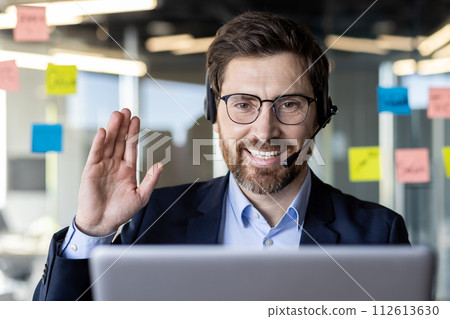 Confident professional businessman wearing a headset and smiling while engaging in a video conference call in a modern office setting. Confident professional businessman wearing a headset and smiling while engaging in a video conference call in a modern office setting. 112613630