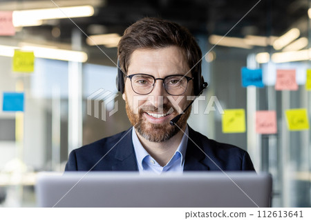A focused businessman in a suit works on a laptop with a headset in a contemporary office environment. A focused businessman in a suit works on a laptop with a headset in a contemporary office environment. 112613641