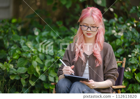 Cute pink hair girl drawing on digital tablet in garden, Woman Doing Freelance Work in Garden, woman with digital tablet with artwork. Cute pink hair girl drawing on digital tablet in garden, Woman Doing Freelance Work in Garden, woman with digital tablet with artwork. 112613820