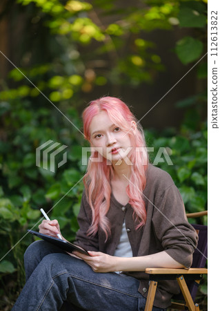 Cute pink hair girl drawing on digital tablet in garden, Woman Doing Freelance Work in Garden, woman with digital tablet with artwork. Cute pink hair girl drawing on digital tablet in garden, Woman Doing Freelance Work in Garden, woman with digital tablet with artwork. 112613822