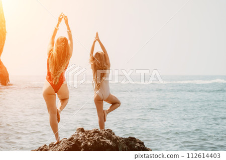Woman and her daughter practicing balancing yoga pose on one leg up together on rock in the sea. Silhouette mother and daughter doing yoga at beach Woman and her daughter practicing balancing yoga pose on one leg up together on rock in the sea. Silhouette mother and daughter doing yoga at beach 112614403