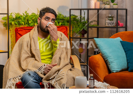 Sick ill Indian man suffering from cold or allergy sits on home chair sneezes wipes snot into napkin Sick ill Indian man suffering from cold or allergy sits on home chair sneezes wipes snot into napkin 112616205