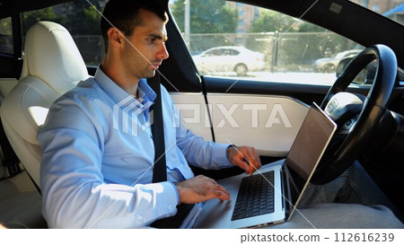 Handsome entrepreneur working on notebook while riding an autonomous self driving electric car at urban road. Male businessman typing text on laptop during riding on electrical vehicle with autopilot 112616239