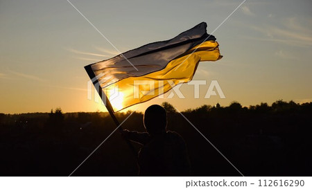 Young man in military uniform waving flag of Ukraine against beautiful sunset at background. Male ukrainian army soldier lifted national banner at countryside. Victory against russian aggression 112616290