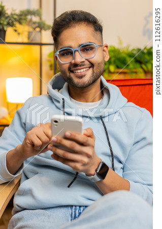 Indian Arabian young man sitting on chair use smartphone share messages on social media application 112616295